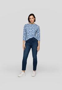 Blau gemusterte Bluse mit Dreiviertelärmeln, kombiniert mit dunkelblauen Jeans und weißen Turnschuhen, stehend vor einem neutralen Hintergrund.