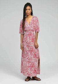 Robe maxi blanche avec motif de gouttes rouges, dotée de manches courtes et d'un col en V, taille cintrée et fente sur le côté. Portée avec des sandales noires.