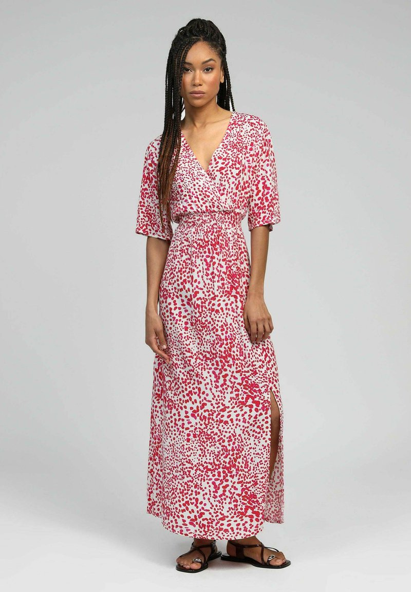 Robe maxi blanche avec motif de gouttes rouges, dotée de manches courtes et d'un col en V, taille cintrée et fente sur le côté. Portée avec des sandales noires.