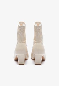 Botas de tobillo blancas hechas de tela suave con un diseño elegante, que cuentan con un tacón grueso y costuras visibles a lo largo de las uniones.