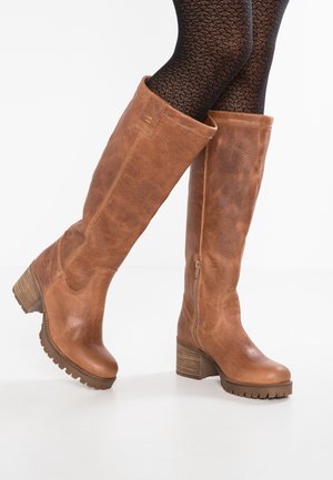 Hoge bruine leren laarzen met blokhakken, gedragen met gestructureerde zwarte panty, waarbij één voet iets omhoog is geheven tegen een witte achtergrond.