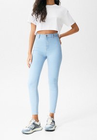 Jeans ajustados de tiro alto en azul claro, confeccionados en denim, combinados con una camiseta blanca cropped y zapatillas gris-azul con detalles en blanco.