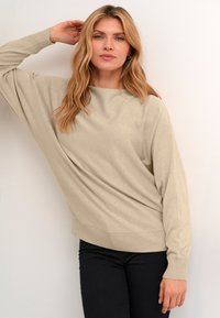 Beige, túlméretezett pulóver, hajónyakú, hosszú ujjú és bordás aljjal. Fekete nadrággal viselve, semleges háttér előtt.