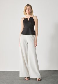 Črni peplum top z V-izrezom in tankimi naramnicami, v kombinaciji z širokimi belimi hlačami. Gladek teksturiran material z minimalističnim dizajnom.