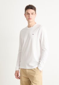 Camiseta blanca de manga larga con cuello redondo, que presenta un pequeño logo en la parte superior izquierda del pecho. El modelo lleva pantalones beige.