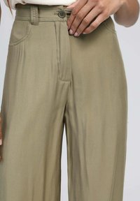 Pantalons larges couleur vert olive en tissu lisse, dotés d'une fermeture à bouton et de poches latérales. Fini texturé avec un léger éclat.