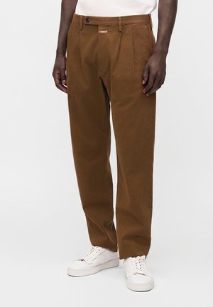 Homme portant un pantalon plissé marron et des baskets blanches, debout les mains détendues le long du corps devant un fond uni.