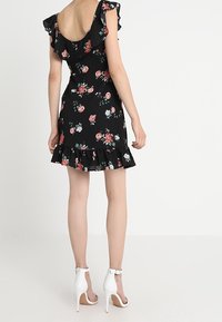 Černé květinové šaty s volánkovými rukávy a lemem, zdobené růžovými a bílými květinovými vzory. Modelka má na sobě bílé sandály na vysokém podpatku.