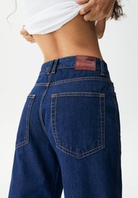 Jeans de mezclilla oscuros de tiro alto con un parche de cuero marrón en la parte trasera, que cuentan con costuras amarillas en contraste y dos bolsillos traseros.