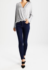 Lys grå indpakningsbluse med lange ærmer, parret med mørkeblå skinny jeans og sorte høje sko. Glat stof, åben halsudskæring.