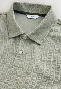 Košeľa s golierom s šedobielym vzorom rybej kosti, dvoma bielymi gombíkmi a štítkom značky "NEXT" vo vnútri goliera.