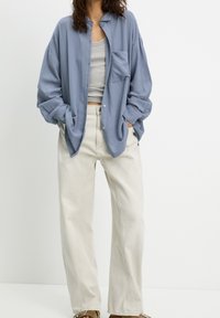 Chemise boutonnée bleu clair à coupe décontractée avec une poche avant, associée à un pantalon large de couleur blanc cassé. Aucun motif ni détail métallique visible.