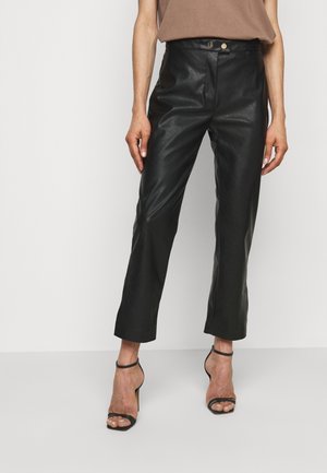 Woman wearing high-waisted black leather pants with a button and zip closure, paired with black ankle-strap high heel sandals.