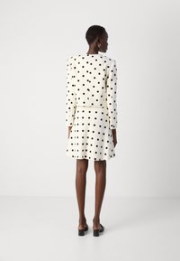 Cream dress with black polka dots, structured jacket, and flared skirt. Features long sleeves and a fitted waist. Smooth, textured fabric.