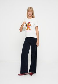 Λευκή μπλούζα με διάγραμμα ενός ανθρωπάκι τζίντζερ και κείμενο, σε συνδυασμό με ναυτικές παντελόνες φαρδιές και κόκκινες γόβες. Το μοντέλο κρατά μια μικρή τσάντα.