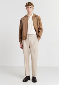 Braune Wildleder-Bomberjacke, cremefarbenes Poloshirt, beige Hose und dunkle Slipper. Glatte Textur und lässiges Design mit taillierter Passform und Bündchen.