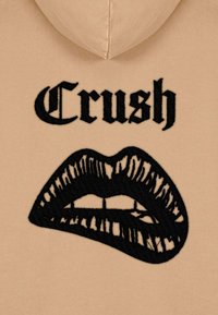 Hoodie beige con un design nero ricamato di labbra e la parola "Crush" in carattere grassetto sopra.