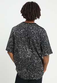 Osoba s krátkými dredy, oblečená v černém tričku se vzorem bílých kapek barvy, viděná zezadu na jednoduchém pozadí.