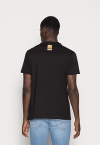 Sort bomulds t-shirt med korte ærmer, der har et farverigt logo mærke bag på nakken. Båret med lyseblå denim jeans.