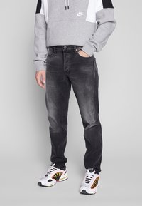 Felpa grigia con un design a blocchi di colore e il logo Nike, abbinata a jeans grigi scuri e sneakers bianche con dettagli rossi e gialli.