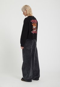 Personne aux cheveux blonds courts portant un pull noir avec un motif tigre coloré dans le dos et un pantalon large gris foncé.