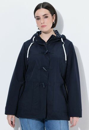 Kobieta o krótkich ciemnych włosach, ubrana w granatową kurtkę z kapturem, zapinaną na drewniane guziki i ozdobioną białymi sznurkami, w połączeniu z niebieskimi dżinsami.