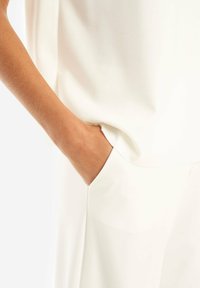 Abbigliamento bianco con una vestibilità rilassata, caratterizzato da una tasca laterale e una texture di tessuto liscia. Una mano è posizionata nella tasca come riferimento.