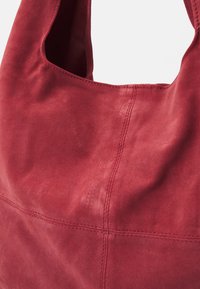 Rote Wildleder-Hobo-Tasche mit einer glatten Textur und sichtbaren Nähten. Das Design umfasst eine geräumige Öffnung und einen gebogenen Schultergurt.
