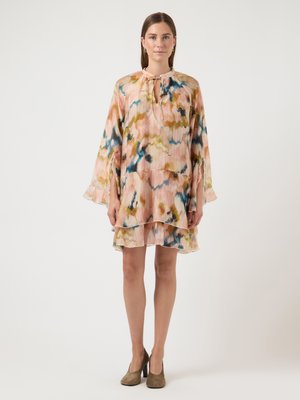 Femme debout portant une robe ample à manches longues, longueur genou, avec des motifs abstraits multicolores et des chaussures à talons beiges.