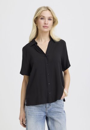 Femeie blondă purtând o cămașă neagră ușor largă, cu mâneci scurte și nasturi, și blugi deschiși la culoare, stând cu o mână în buzunar.