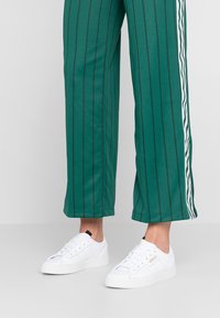 Gröna randiga byxor med lös passform och vita sidodetaljer, i kombination med vita sneakers med svarta och guldiga detaljer.