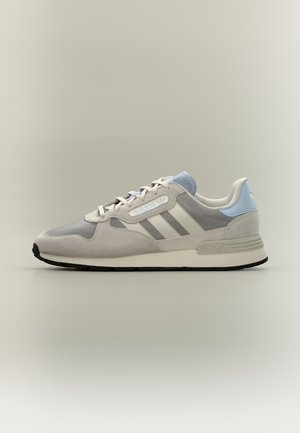 Ανοιχτό γκρι αθλητικό παπούτσι Adidas με λευκές ρίγες, ανοιχτά μπλε λεπτομέρειες, κορδόνια και μαύρη σόλα, εμφανίζεται από το πλάι σε ουδέτερο φόντο.