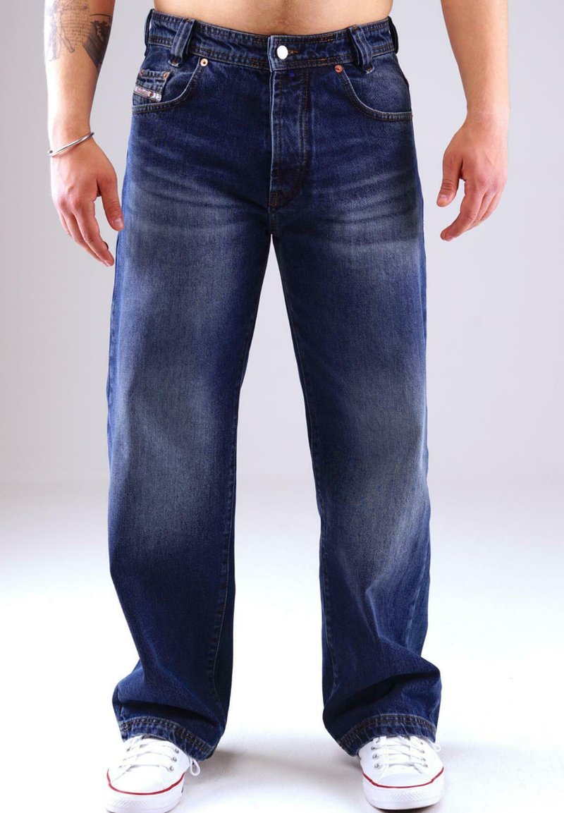 Mann in lässigen, dunkelblauen Jeans und weißen Sneakers, der vor einem schlichten, hellen Hintergrund steht und Unterkörper sowie Beine zeigt.