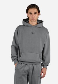 Young man with medium-length brown hair wearing a gray hoodie and matching sweatpants with hands in pockets against a plain background.
