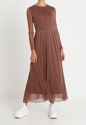 Femme portant une robe longue à manches longues, marron scintillante, associée à des baskets blanches épaisses, debout devant un fond uni.