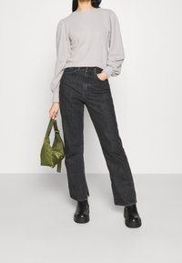 Šedý svetr s balónovými rukávy, černé džíny s vysokým pasem a černé platformové boty. Drží zelenou saténovou tašku. Jednoduchý, čistý design.