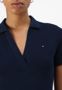 Prim-plan cu o persoană care poartă o cămașă albastru închis cu guler, cu un mic logo roșu, alb și albastru pe piept și cercei aurii rotunzi.