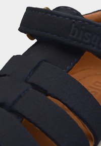 Gros plan sur les brides en cuir bleu marine d'une sandale avec des coutures visibles et une semelle intérieure en cuir beige en dessous.