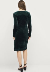 Dunkelgrünes Samtkleid mit langen Ärmeln, taillierter Silhouette und knielangem Saum. Verfügt über einen subtilen Glanz und eine glatte Textur, kombiniert mit schwarzen Absätzen.