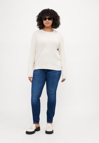 Lys beige genser med teksturert finish, lange ermer og rund hals, parret med middels blå jeans og lukkede sko. Svarte solbriller brukt.