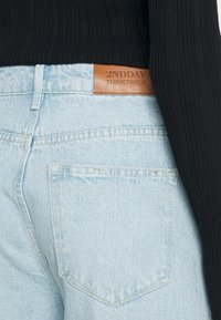 Vaqueros de denim azul claro con una superficie texturizada, que presentan una etiqueta de cuero que dice "2NDDAY THINKTWICE" en la cintura y en un bolsillo trasero.