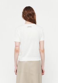Witte kortärmelige top gemaakt van katoen met een eenvoudige structuur. Heeft een klein logopatch op de bovenrug. Gecombineerd met een beige rok.
