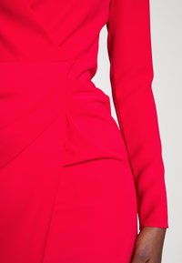 Närbild av en person som bär ett livfullt röd, långärmad plagg med samlad tygdetalj vid midjan.