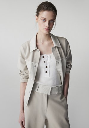 Młoda kobieta o jasnych oczach i zaczesanych do tyłu włosach, ubrana w beżową kurtkę, zapinaną na guziki białą koszulkę na ramiączkach i beżowe spodnie z wysokim stanem oraz paskiem.