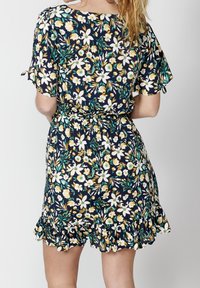 Žena oblečená v námořnickém modrém květinovém šatech s bílými a žlutými květinami, krátké rukávy, elastický pas a volánkový lem, zobrazená zezadu.