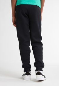 Sorte bomulds joggerbukser med elastiske manchetter, parret med hvide sneakers med sorte accenter. Bagfra vises pasform og design.