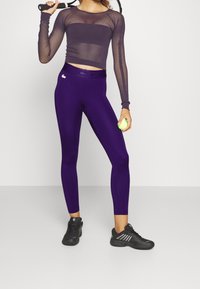 Leggins morados y una camiseta de manga larga de malla, sosteniendo una pelota de tenis, con zapatillas deportivas negras. Diseño simple, silueta ajustada, material transpirable.