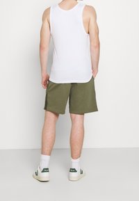 Muž v bílém tílku, olivově zelených kraťasech ke kolenům, bílých ponožkách a bílých teniskách se zeleným logem, stojící na pozadí s čistě bílou barvou.