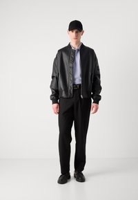 Black leather bomber jacket with ribbed cuffs and a zippered front, worn over a light blue polo, paired with black trousers and black shoes.