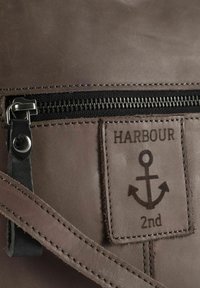 Bruine leren tas met zwarte ritssluiting, voorzien van een textuurtag met geperst opschrift "HARBOUR" en een ankersymbool.